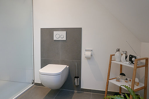 Nach Sanierung: Freistehende Toilette vor abgesetzten grauen Fliesenspiegel.